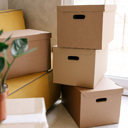 MovingStacked cardboard boxes beside a yellow sofa and a potted plant, in a bright room with natural light, suggesting moving or organizing.