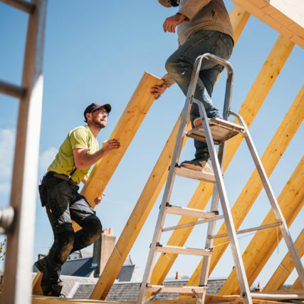 ContractorTwo construction workers assemble a wooden roof frame. One stands on a ladder, the other hands him a beam. The scene conveys teamwork under a clear blue sky.