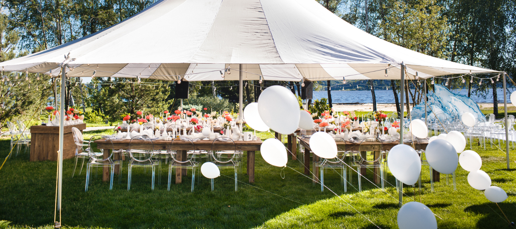 Outdoor wedding reception under a white tent by a lake. Tables are set with vibrant floral arrangements and surrounded by clear chairs. White balloons float nearby.